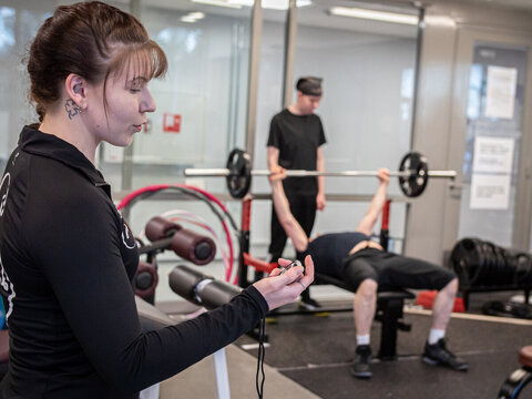 Liikunnanohjaaja Sirke Savolainen ottaa aikaa, kun ryhmän osallistujat harjoittelevat lihaskuntoa painotangolla.